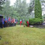 Grupa ludzi z flagami stoi na trawniku, w tle drzewa. Obok flagi Polski, widoczny jest pomnik z krzyżem. Uroczystość odbywa się w deszczu.
