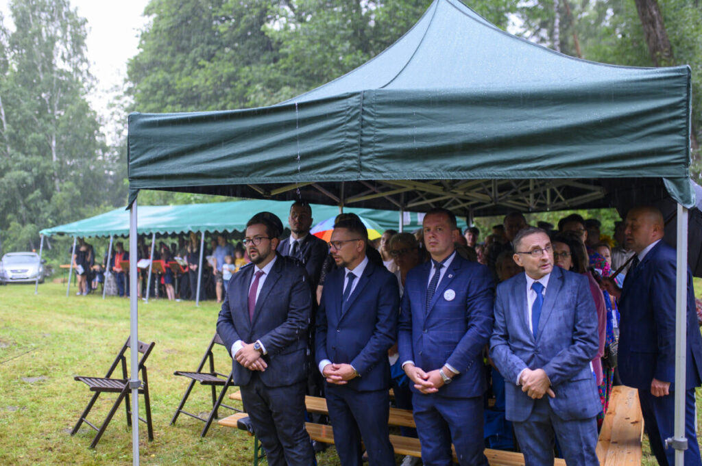 Grupa osób w garniturach stoi pod zielonym namiotem. Pada deszcz, a w tle widać zielone drzewa i kolejny namiot z ludźmi. Po lewej stoi kilka pustych krzeseł na trawie.