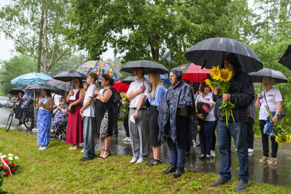 Grupa ludzi stoi pod parasolami na trawie, ubrani w jasne i ciemne kolory. W tle drzewa, a osoba trzyma słoneczniki. Pada deszcz.