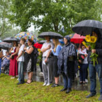 Grupa ludzi stoi pod parasolami na trawie, ubrani w jasne i ciemne kolory. W tle drzewa, a osoba trzyma słoneczniki. Pada deszcz.