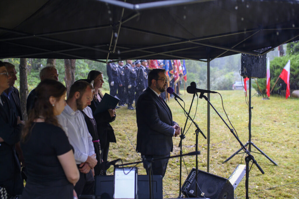 Pod namiotem grupa ludzi uczestniczy w ceremonii przy mikrofonie. W tle widoczne są drzewa i flagi Polski. Pada deszcz.