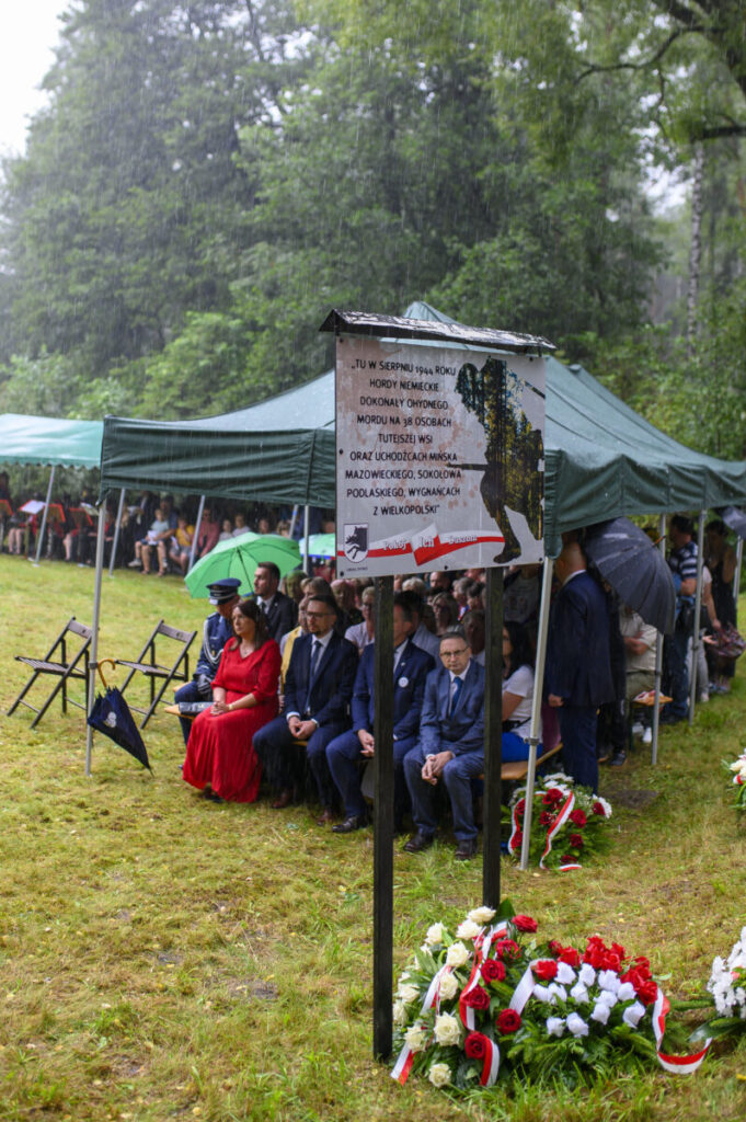 Obraz przedstawia uroczystość plenerową pod zielonymi namiotami w deszczu. Ludzie siedzą na krzesłach na trawie, z kwiatami wokół. W centrum widnieje tablica upamiętniająca historyczne wydarzenie.