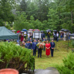 Zdjęcie przedstawia uroczystość na świeżym powietrzu. Grupa ludzi z wieńcami stoi na zielonej trawie. W tle są drzewa i namioty, a uczestnicy trzymają parasole.