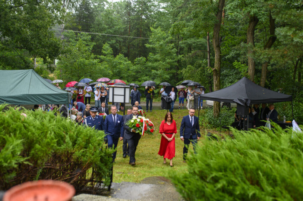 Grupa ludzi z kwiatami na pewno w uroczystości plenerowej. Zielona trawa, drzewa i namioty w tle. Parasole w różnych kolorach chronią przed deszczem.