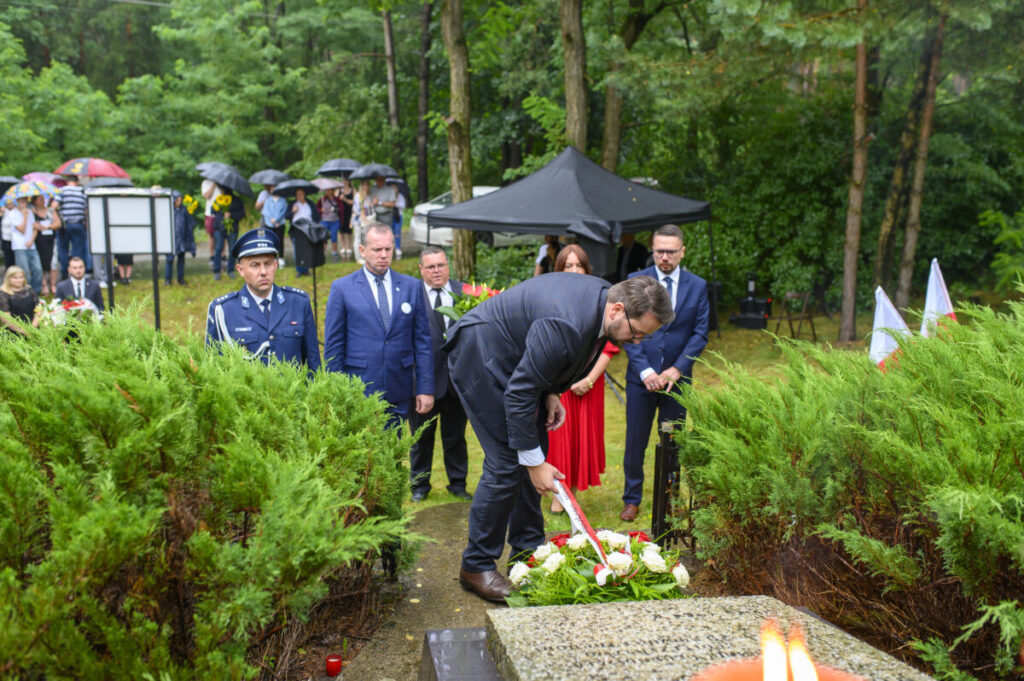 Na zdjęciu ludzie składają kwiaty na grobie w zielonym lesie. Towarzyszy im mężczyzna w mundurze, a w tle są widoczne parasole. Atmosfera uroczysta, tło pełne zieleni.