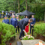 Na zdjęciu ludzie składają kwiaty na grobie w zielonym lesie. Towarzyszy im mężczyzna w mundurze, a w tle są widoczne parasole. Atmosfera uroczysta, tło pełne zieleni.