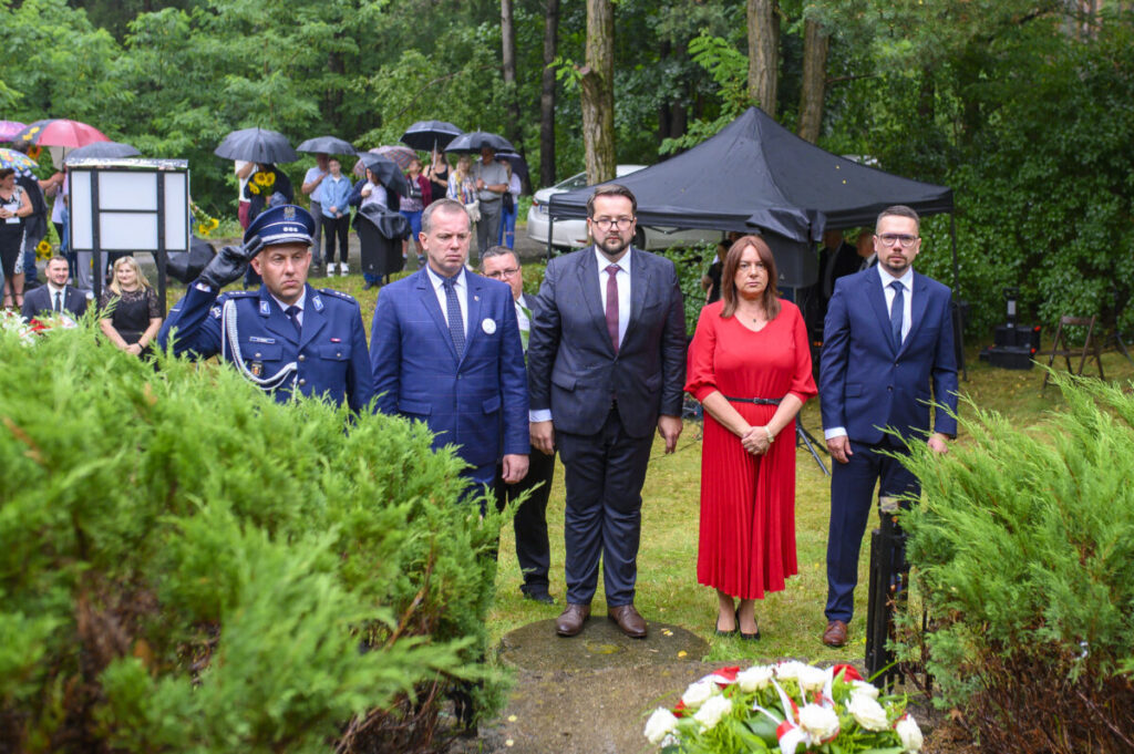 Grupa ludzi stoi na trawniku podczas uroczystości w parku. Mają na sobie eleganckie ubrania; jest deszczowo, widoczne parasole. W tle gęsta zieleń i namiot.