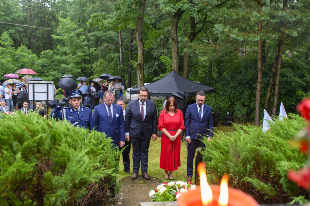 Pięć osób w eleganckich strojach stoi w lesie, obok złożonych kwiatów. Tło: zielone drzewa, ludzie z parasolkami. W tle czarny namiot.