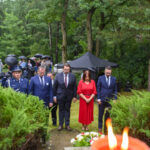 Pięć osób w eleganckich strojach stoi w lesie, obok złożonych kwiatów. Tło: zielone drzewa, ludzie z parasolkami. W tle czarny namiot.