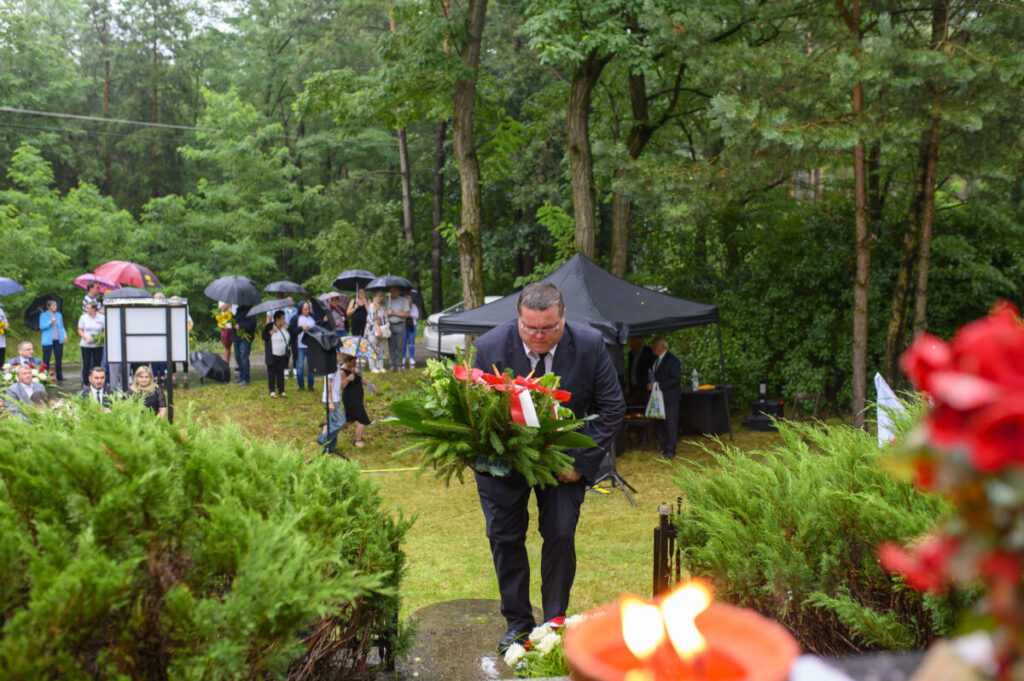 Mężczyzna w czarnym garniturze składa wieniec na tle zielonego lasu. Grupa osób z parasolami w tle. Czerwone kwiaty z przodu.