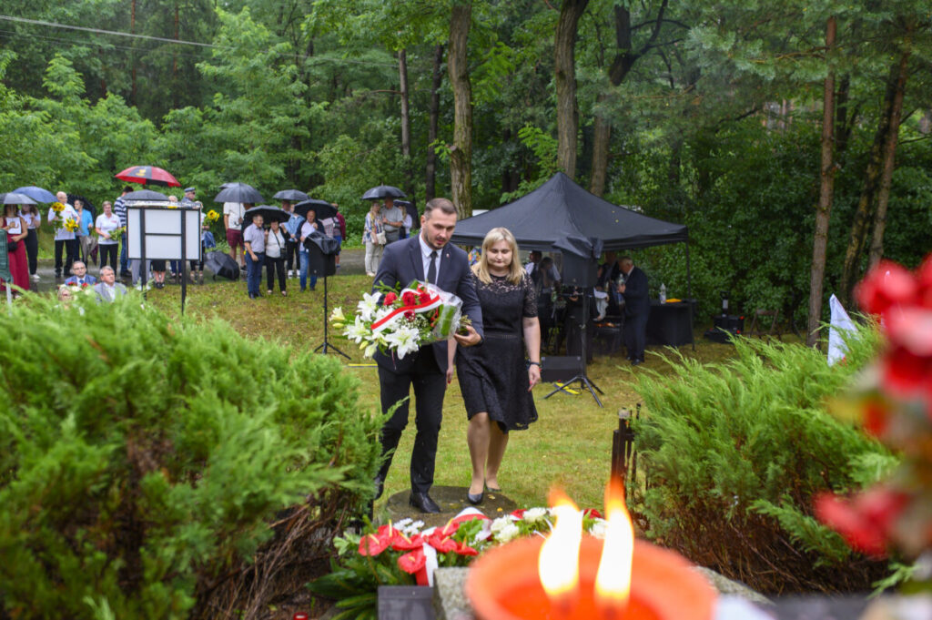 Dwójka ludzi składa wiązankę kwiatów przed pomnikiem. W tle zielony las, uczestnicy z parasolkami. Pierwszy plan to płomień świecy.