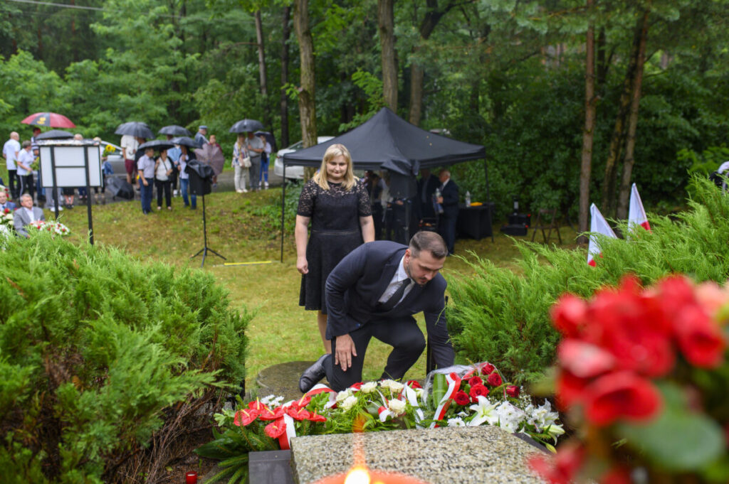 Mężczyzna i kobieta składają kwiaty na grobie, w tle zielone drzewa. Widać czarne parasole i czerwone kwiaty. Tło stanowi las.