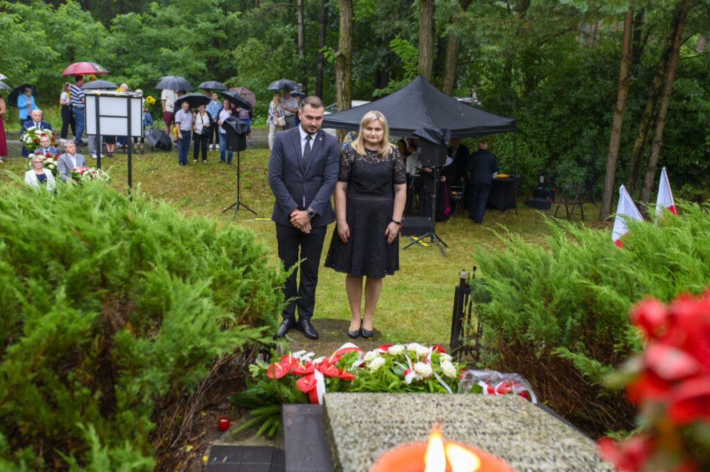 Dwóch ludzi w czerni stoi przed pomnikiem z kwiatami. Tło to zielone drzewa, mokra trawa i zgromadzony tłum z parasolami. U dołu płonie świeca.