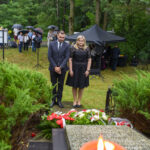 Dwóch ludzi w czerni stoi przed pomnikiem z kwiatami. Tło to zielone drzewa, mokra trawa i zgromadzony tłum z parasolami. U dołu płonie świeca.