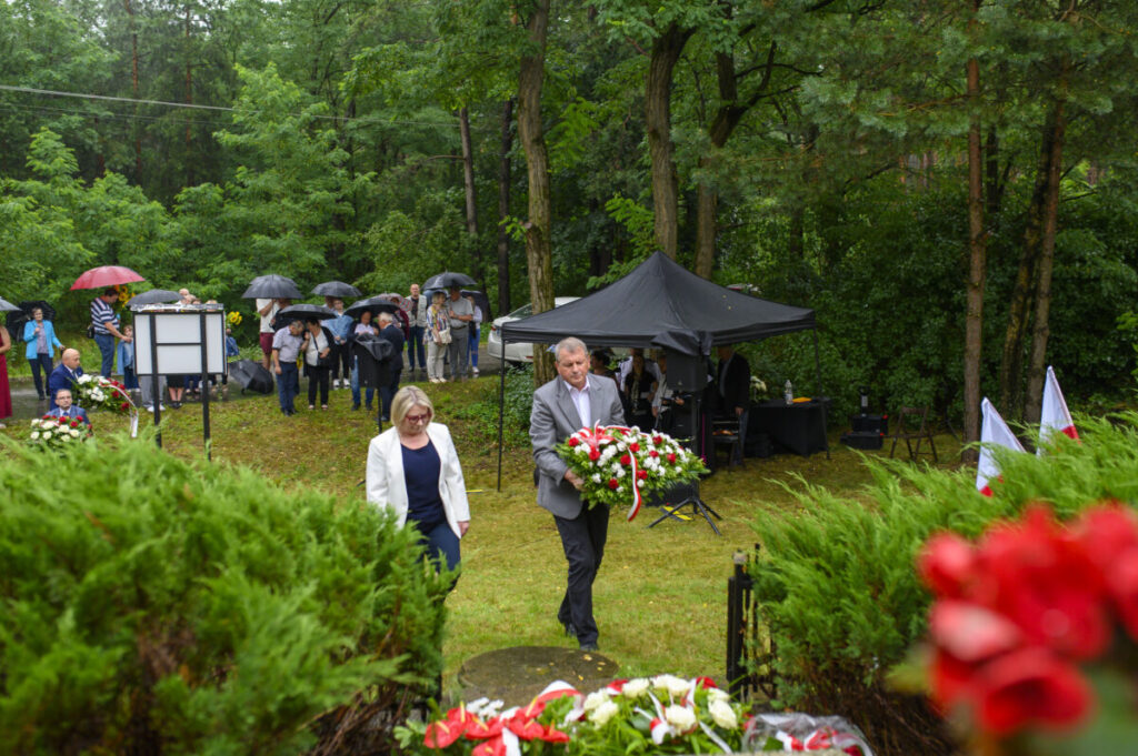 Na zdjęciu widoczni są ludzie pod parasolami w lesie podczas ceremonii. Mężczyzna niesie wieniec z biało-czerwonymi kwiatami. W tle namiot i drzewa.
