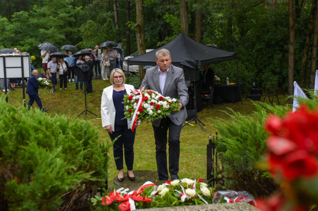 Para składa wieniec na tle zieleni. Ludzie z parasolami stoją w oddali. Czarny namiot widoczny po prawej stronie.
