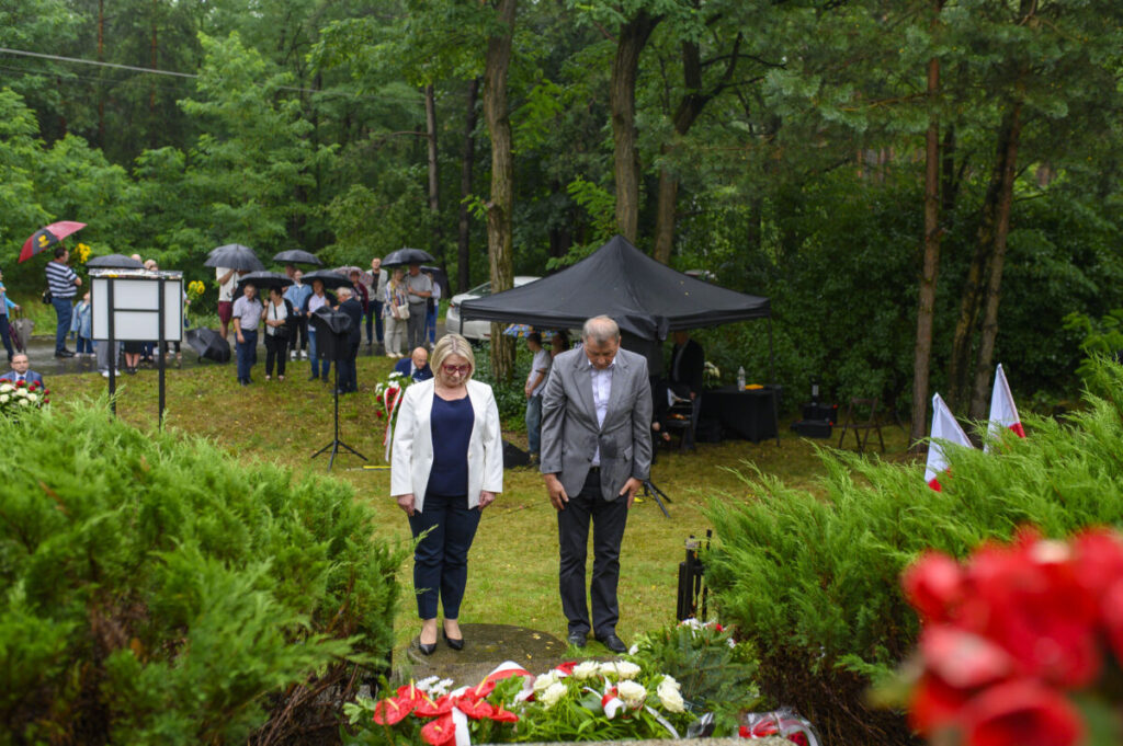 Dwóch ludzi w eleganckich ubraniach składa hołd przy wieńcach. W tle las i ludzie z parasolami. Scena odbywa się na zewnątrz.