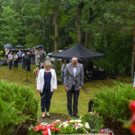 Dwóch ludzi w eleganckich ubraniach składa hołd przy wieńcach. W tle las i ludzie z parasolami. Scena odbywa się na zewnątrz.