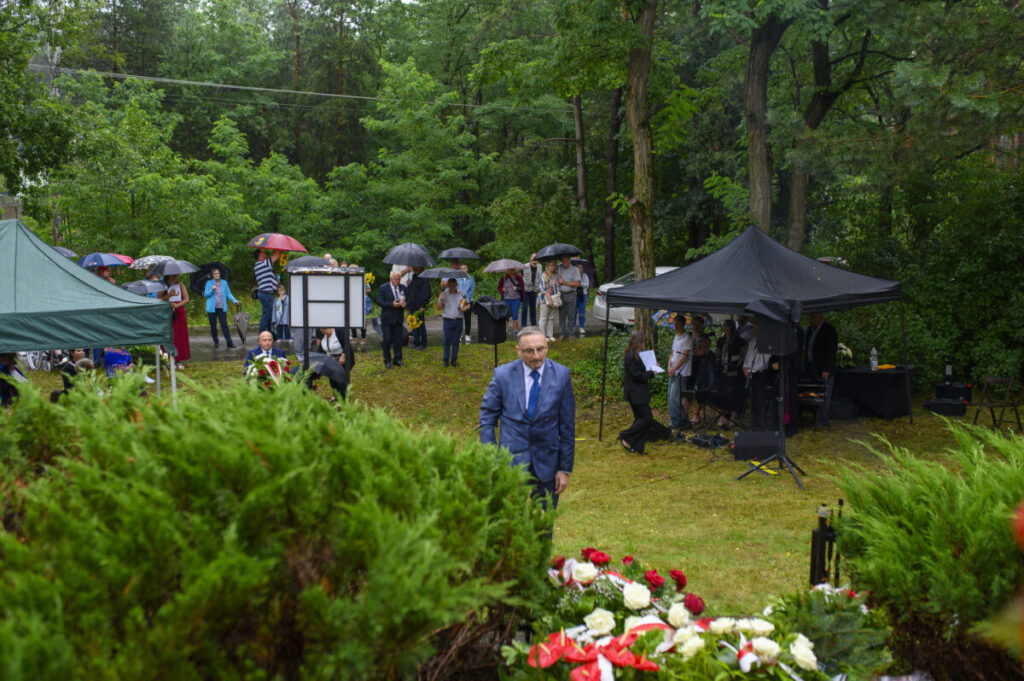 Grupa ludzi z parasolkami stoi na trawie wśród drzew. Na pierwszym planie kwiaty, z tyłu zielony i czarny namiot. Tło to bujna zieleń i deszczowa atmosfera.