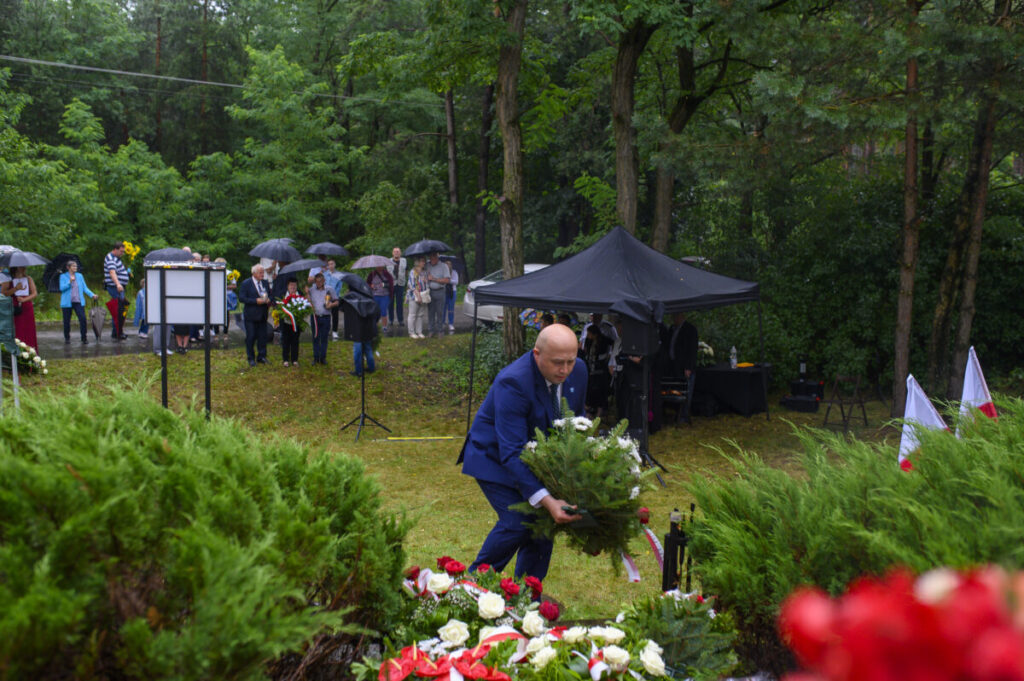 Na zdjęciu mężczyzna w niebieskim garniturze składa wieniec przy pomniku. W tle ludzie z parasolami stoją w lesie. Otoczenie jest zielone z namiotem.