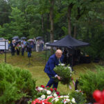 Na zdjęciu mężczyzna w niebieskim garniturze składa wieniec przy pomniku. W tle ludzie z parasolami stoją w lesie. Otoczenie jest zielone z namiotem.
