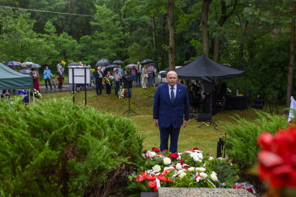Mężczyzna w garniturze stoi przed czerwono-białymi kwiatami. W tle widoczne są drzewa, ludzie z parasolami i czarne namioty. Dominuje zielona sceneria.