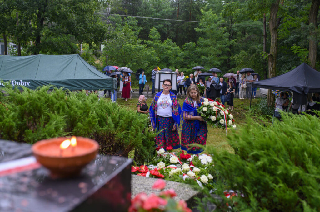 Na zdjęciu dwie kobiety w kolorowych strojach niosą wieniec. W tle ludzie pod parasolami, drzewa. Ciepłe światło świecy na pierwszym planie.