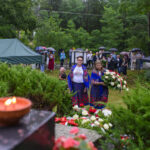 Na zdjęciu dwie kobiety w kolorowych strojach niosą wieniec. W tle ludzie pod parasolami, drzewa. Ciepłe światło świecy na pierwszym planie.