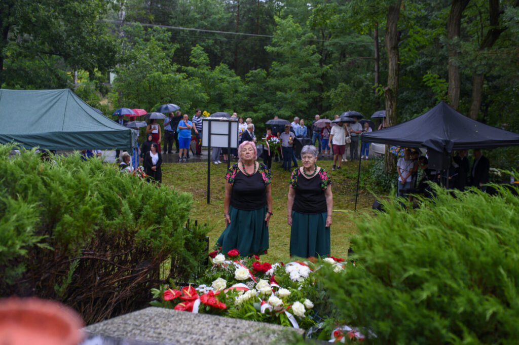 Dwie starsze kobiety w ludowych strojach stoją przed wieńcami. W tle zebrani ludzie z parasolami. Scena na tle zieleni.