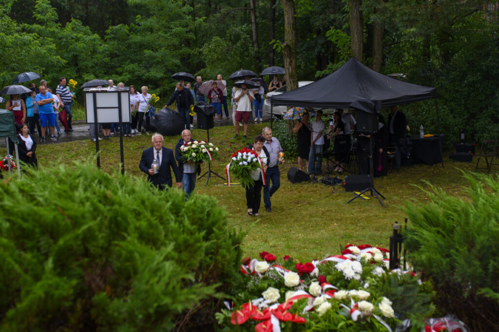 Grupa ludzi z kwiatami i parasolami stoi na trawniku podczas deszczu. Na pierwszym planie widać wieńce kwiatowe w barwach narodowych. W tle zielone drzewa i harcerze pod namiotem.