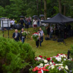 Grupa ludzi z kwiatami i parasolami stoi na trawniku podczas deszczu. Na pierwszym planie widać wieńce kwiatowe w barwach narodowych. W tle zielone drzewa i harcerze pod namiotem.