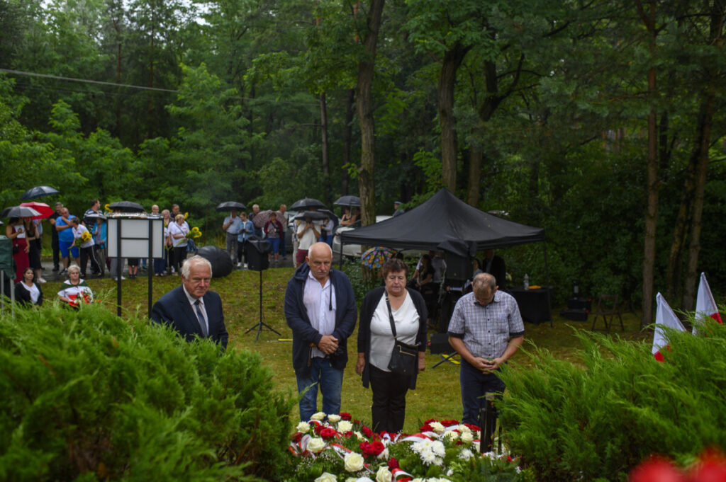 Grupa ludzi stoi na uroczystości w lesie, niektórzy trzymają parasole. Przed nimi kwiaty w barwach czerwono-białych. W tle namiot i drzewa.