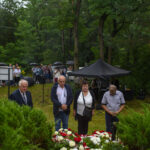Grupa ludzi stoi na uroczystości w lesie, niektórzy trzymają parasole. Przed nimi kwiaty w barwach czerwono-białych. W tle namiot i drzewa.