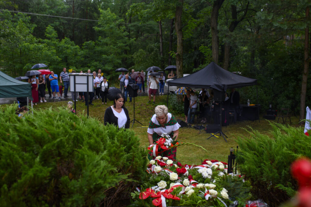 Ludzie składają wieńce w zielonym parku. W tle namioty i tłum pod parasolami. Atmosfera deszczowa, dominują zielenie i czerwienie kwiatów.