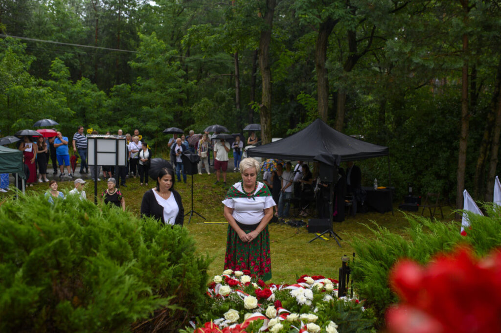 Grupa ludzi z parasolami na uroczystości w lesie; z przodu kompozycja kwiatów czerwono-białych; w tle namiot i drzewa.