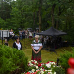 Grupa ludzi z parasolami na uroczystości w lesie; z przodu kompozycja kwiatów czerwono-białych; w tle namiot i drzewa.