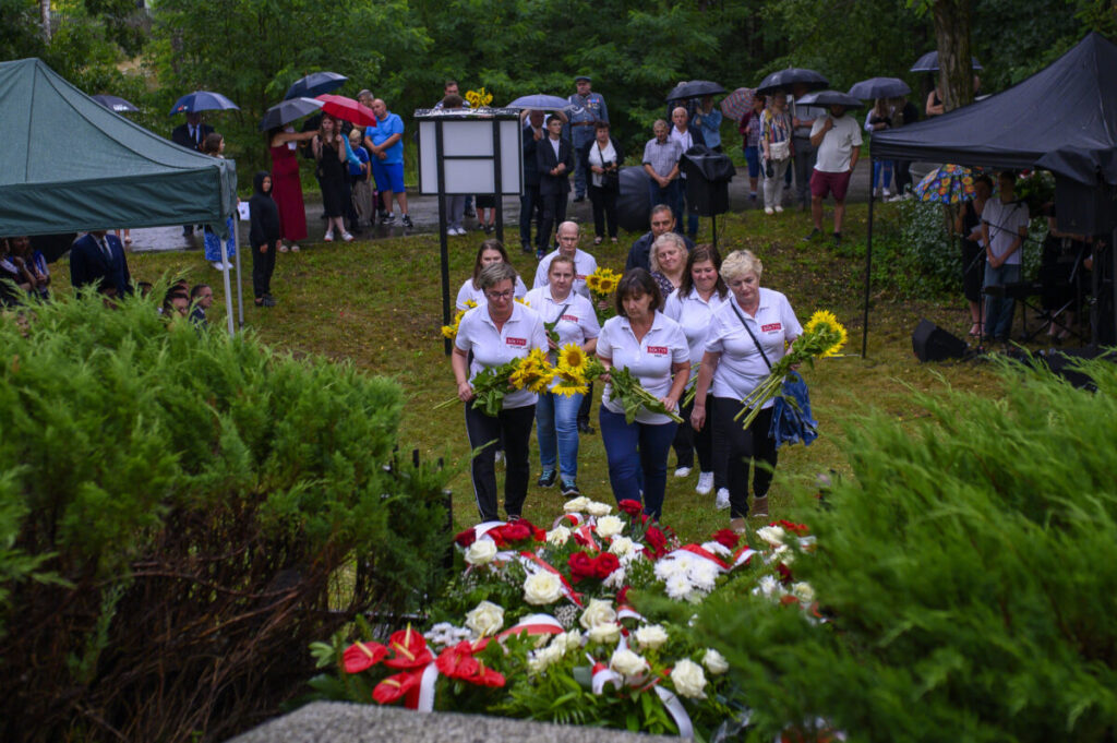 Grupa osób w białych koszulkach z kwiatami stoi przed wieńcami. Tło zielone, ludzi osłaniają kolorowe parasole. Atmosfera poważna.