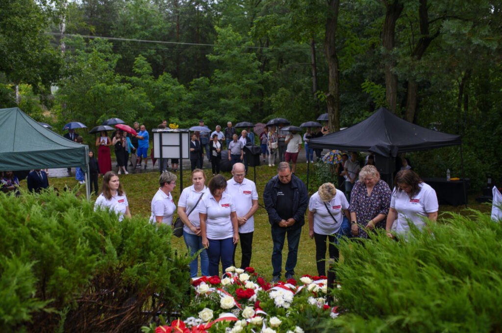 Grupa ludzi w białych koszulkach pochyla się nad kwiatami. W tle inni ludzie trzymają parasole. Wokół zielone drzewa i namioty.