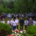 Grupa ludzi w białych koszulkach pochyla się nad kwiatami. W tle inni ludzie trzymają parasole. Wokół zielone drzewa i namioty.