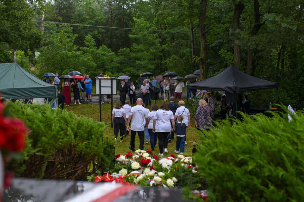 Grupa ludzi pod parasolami na tle zielonego lasu. Namioty w ciemnych kolorach. Kwiaty w jasnych barwach na pierwszym planie.