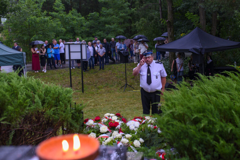 Mężczyzna w białej koszuli salutuje obok kwiatów. W tle ludzi pod parasolami i zielone drzewa. Atmosfera uroczysta, mokre od deszczu.