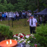 Mężczyzna w białej koszuli salutuje obok kwiatów. W tle ludzi pod parasolami i zielone drzewa. Atmosfera uroczysta, mokre od deszczu.