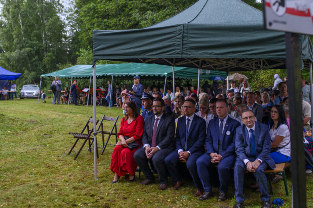 Na zdjęciu widać ludzi siedzących pod zielonym namiotem na trawie. Mężczyźni w garniturach, kobieta w czerwonej sukni. W tle drzewa i niebieski namiot.