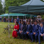 Na zdjęciu widać ludzi siedzących pod zielonym namiotem na trawie. Mężczyźni w garniturach, kobieta w czerwonej sukni. W tle drzewa i niebieski namiot.