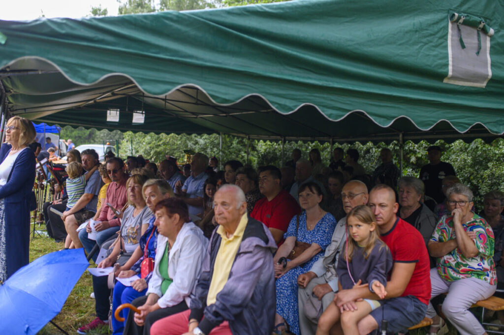 Grupa osób siedzi pod zielonym namiotem na ławach. Tło składa się z zieleni drzew. Przed ludźmi leży niebieski parasol.