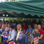 Grupa osób siedzi pod zielonym namiotem na ławach. Tło składa się z zieleni drzew. Przed ludźmi leży niebieski parasol.