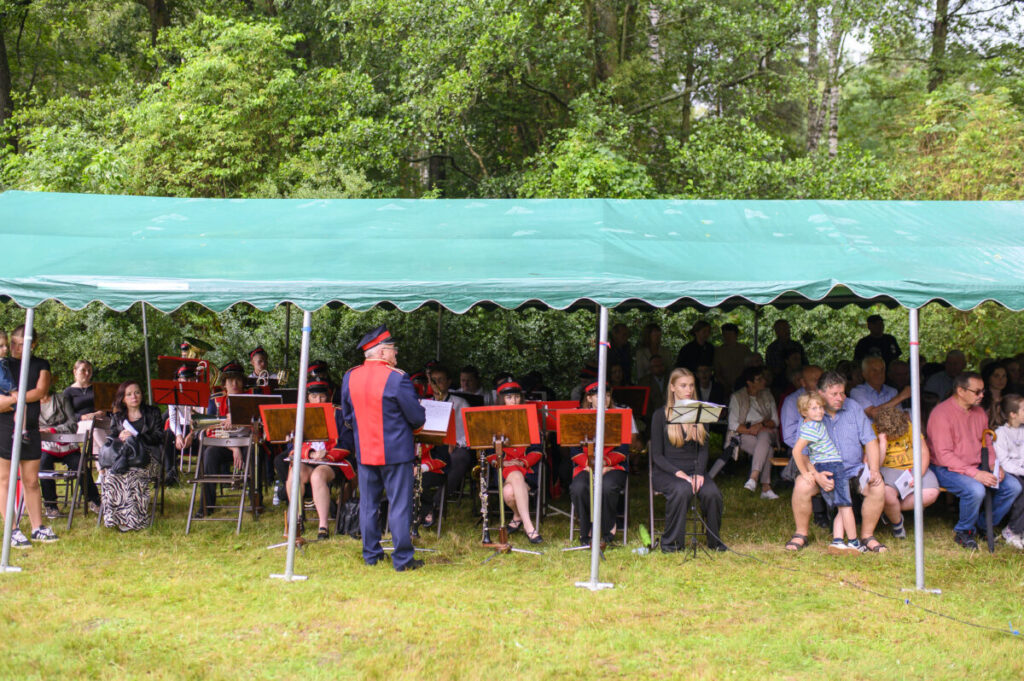 Pod zielonym namiotem orkiestra siedzi na krzesłach, ubrana w czerwone mundury. W tle zielone drzewa. Obok widownia.