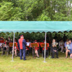 Pod zielonym namiotem orkiestra siedzi na krzesłach, ubrana w czerwone mundury. W tle zielone drzewa. Obok widownia.