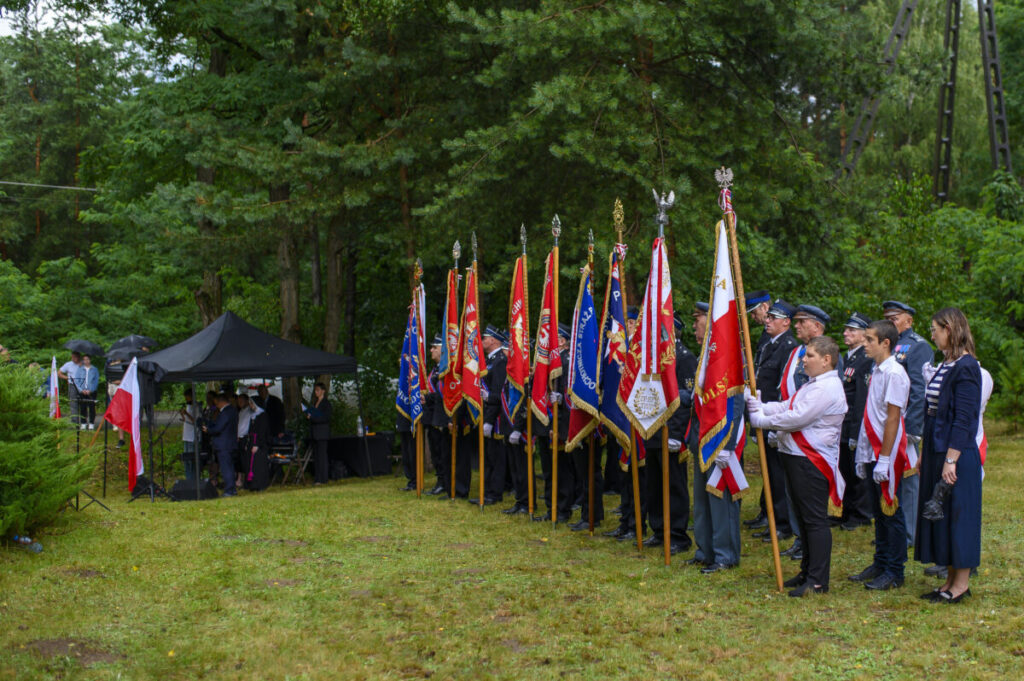 Grupa osób w paradnych strojach trzyma kolorowe sztandary na tle zielonego lasu. Obok czarny namiot i mężczyźni w mundurach. Trawa jest wilgotna, a niebo pochmurne.