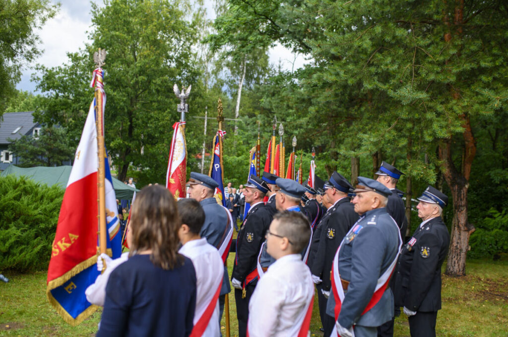 Grupa osób w mundurach trzyma sztandary w parku. Ubrani w ciemne garnitury, otoczeni zielenią drzew. Czerwono-białe flagi dominują.